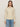 A person stands against a plain light wall, wearing Sanctuary Clothing's "one in a million cable crewneck sweater" in vanilla, layered over a ruffled white top with blue jeans. They have straight, shoulder-length brown hair and a neutral expression.