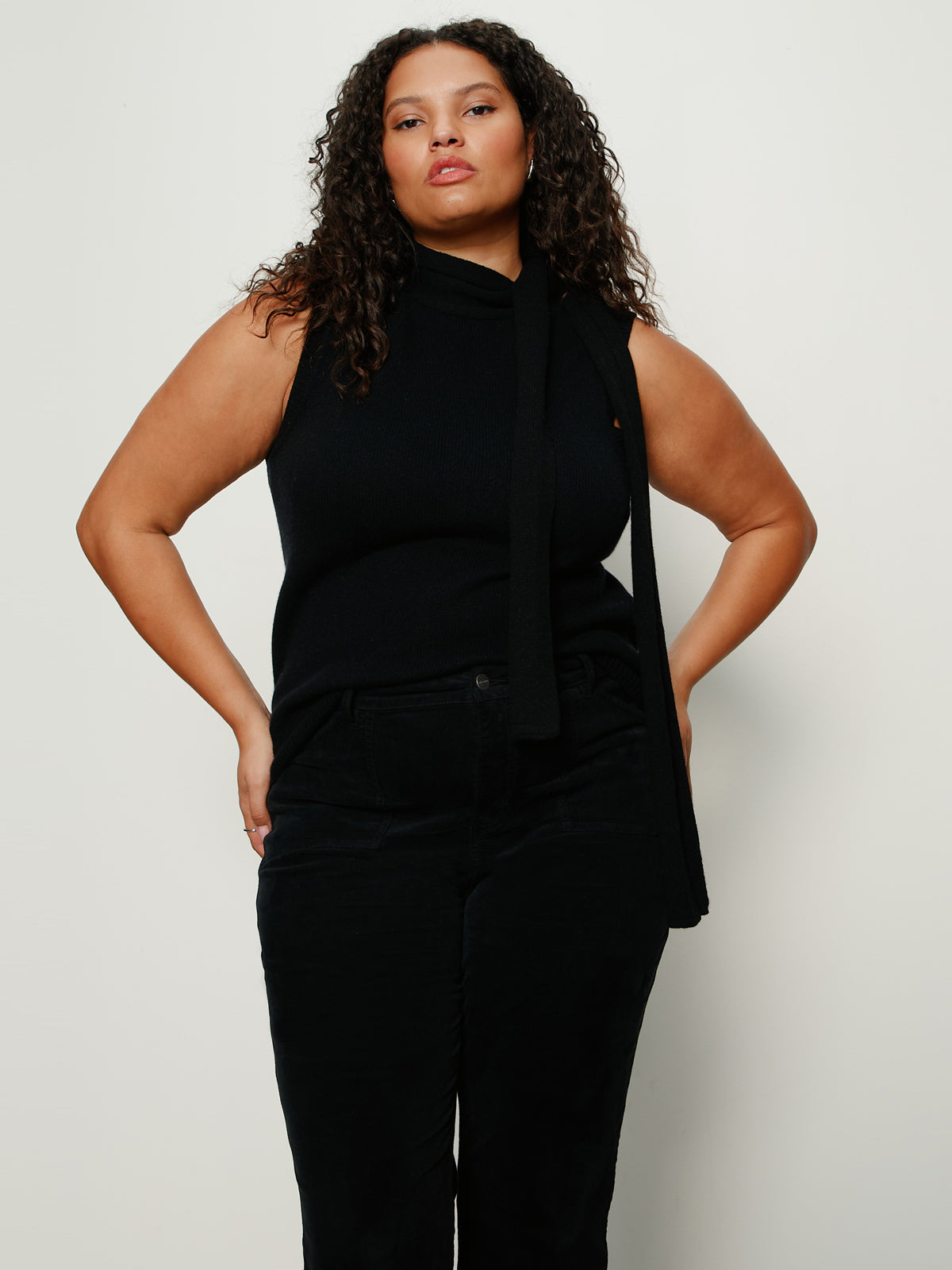 A woman with curly hair wears the Sanctuary Clothing scarf sweater shell in black extended sizing, paired with black pants, standing against a plain white background with her hands on her hips.