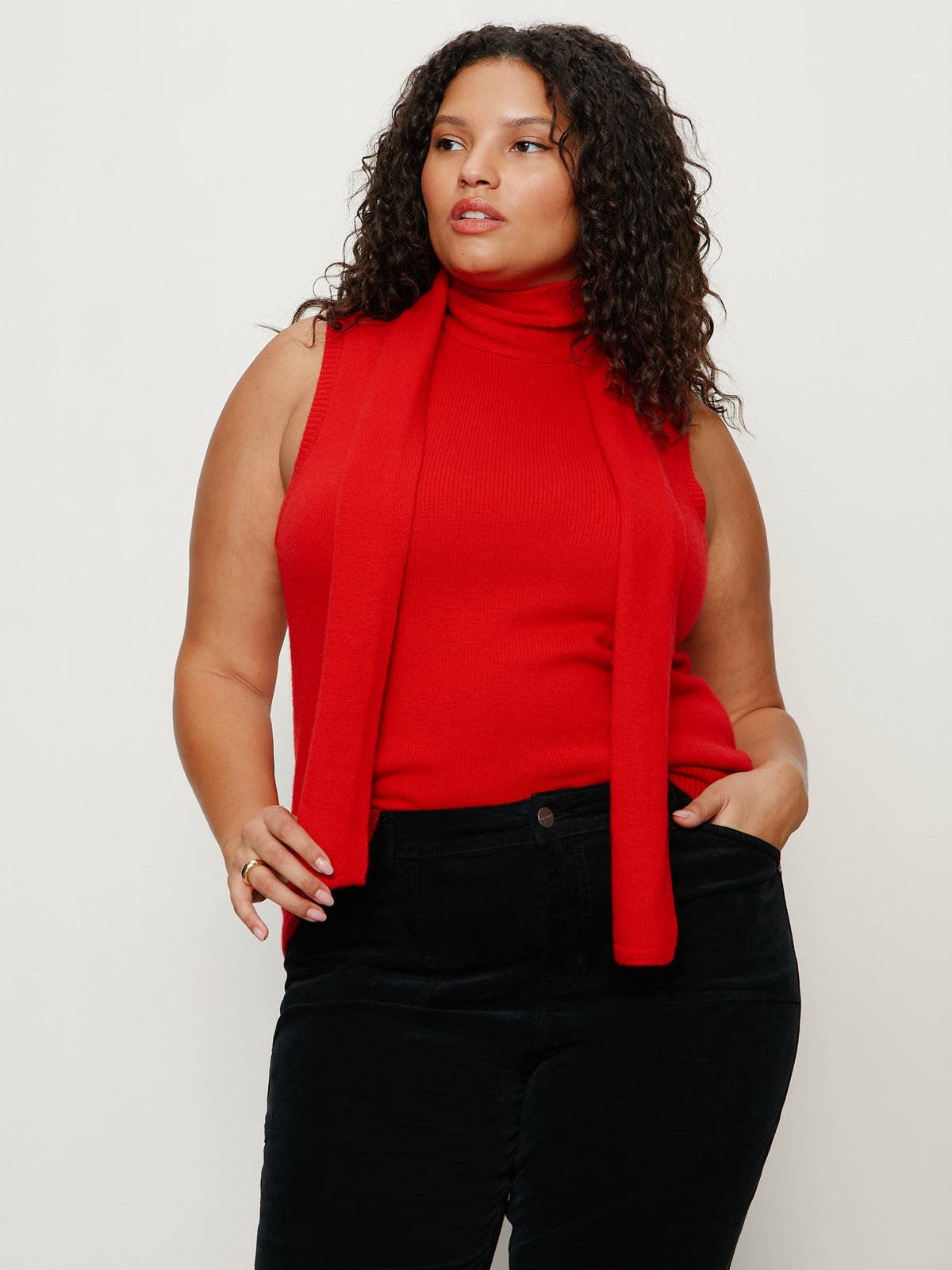 A woman with curly brown hair wears Sanctuary Clothing's scarf sweater shell in cherry lacquer extended sizing, paired with black pants, standing against a plain light background with one hand in her pocket.