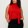A woman with curly brown hair wears Sanctuary Clothing's scarf sweater shell in cherry lacquer extended sizing, paired with black pants, standing against a plain light background with one hand in her pocket.
