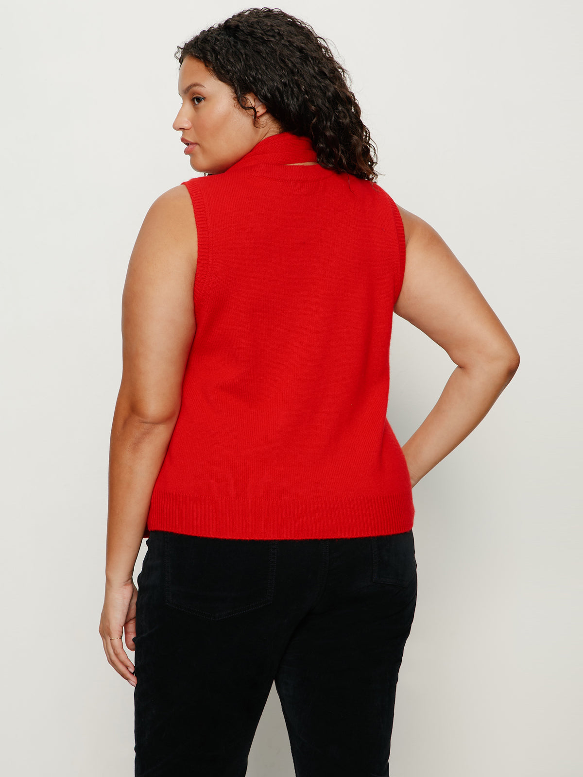A woman with curly dark hair models the Sanctuary Clothing scarf sweater shell in cherry lacquer (extended sizing), a sleeveless red turtleneck, paired with black pants, standing with her back to the camera against a light background.