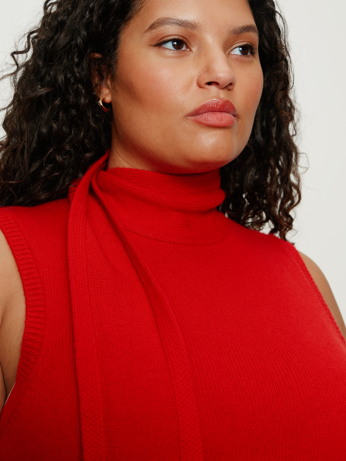 A woman with curly dark hair wears the Sanctuary Clothing scarf sweater shell in cherry lacquer, available in extended sizing. She stands confidently, looking to the side against a plain light background.