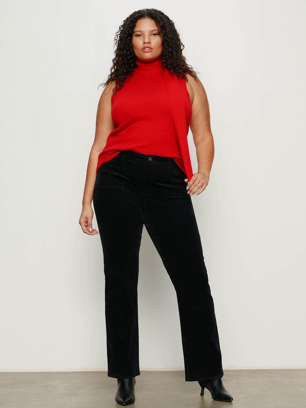 A woman with curly hair models the Sanctuary Clothing scarf sweater shell in cherry lacquer (extended sizing), styled with black pants and ankle boots. She stands against a plain, light background, gazing confidently at the camera.