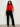 A woman with curly hair models the Sanctuary Clothing scarf sweater shell in cherry lacquer (extended sizing), styled with black pants and ankle boots. She stands against a plain, light background, gazing confidently at the camera.