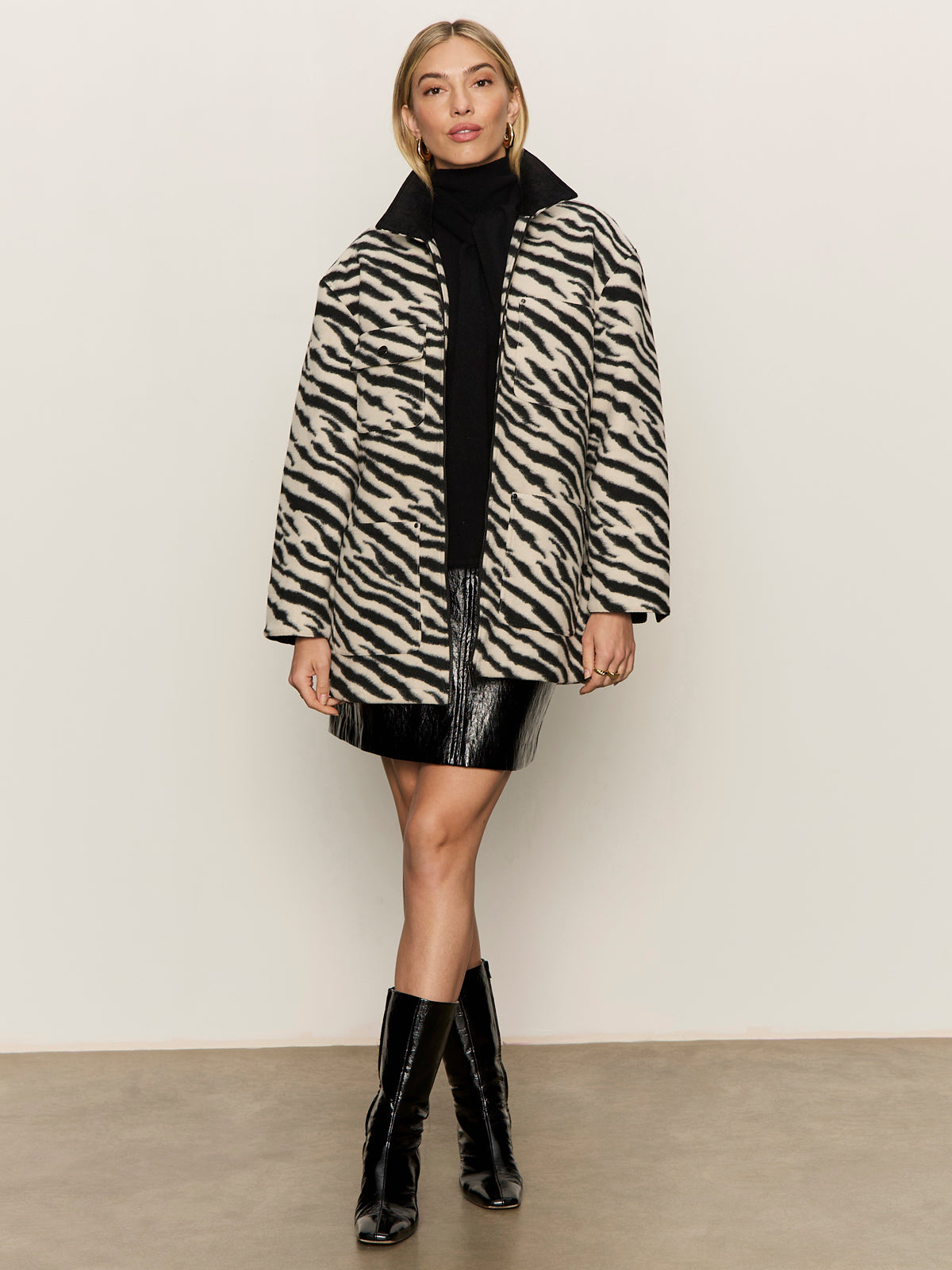 A woman with blonde hair styled back wears the Sanctuary Clothing scarf sweater shell in black, paired with a short black skirt and shiny knee-high boots, standing against a plain background with a neutral expression.