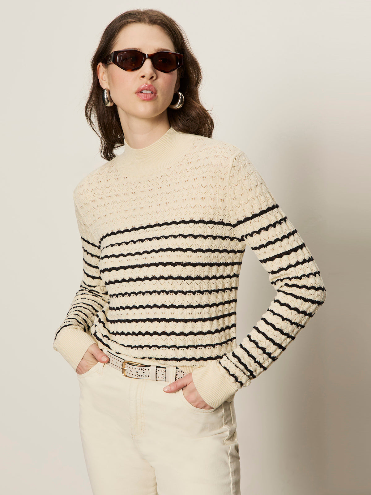 A woman stands against a plain light background wearing Sanctuary Clothing’s forever striped mock neck sweater in vanilla creme with black stripes, paired with sunglasses, hoop earrings, and ivory pants, hands in her pockets.