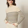 A woman stands against a plain light background wearing Sanctuary Clothing’s forever striped mock neck sweater in vanilla creme with black stripes, paired with sunglasses, hoop earrings, and ivory pants, hands in her pockets.