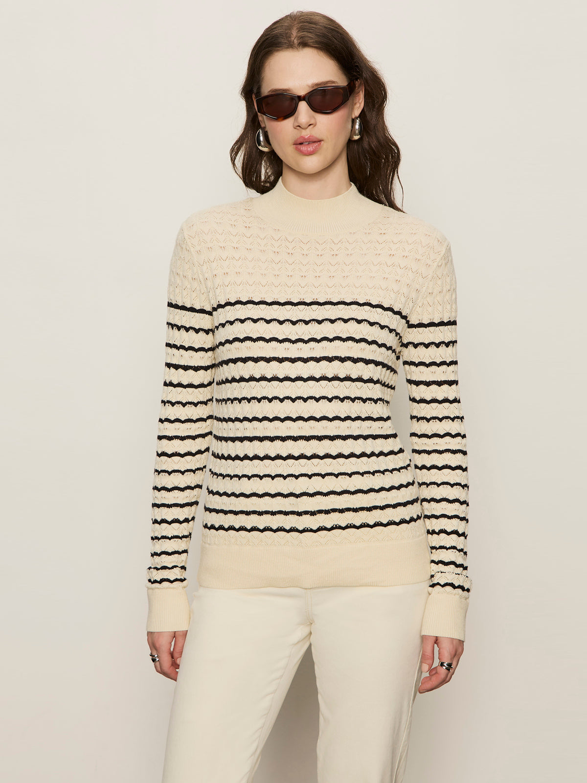 A woman in the Sanctuary Clothing forever striped mock neck sweater in vanilla creme with black stripes, paired with light pants and gold hoop earrings, stands against a plain background wearing dark sunglasses.