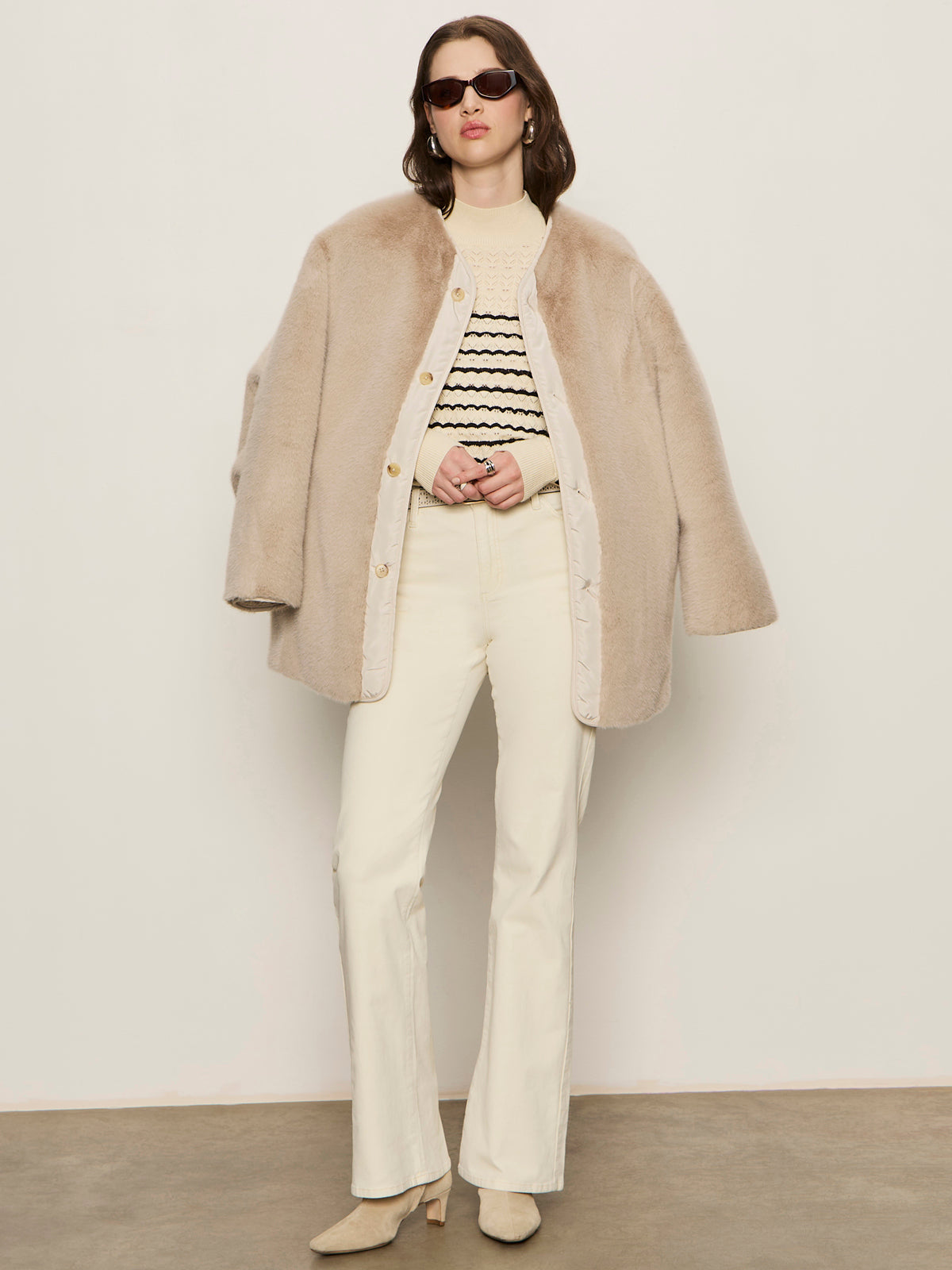 A woman stands against a plain wall wearing sunglasses, a beige faux fur coat, a cream and black striped sweater, off