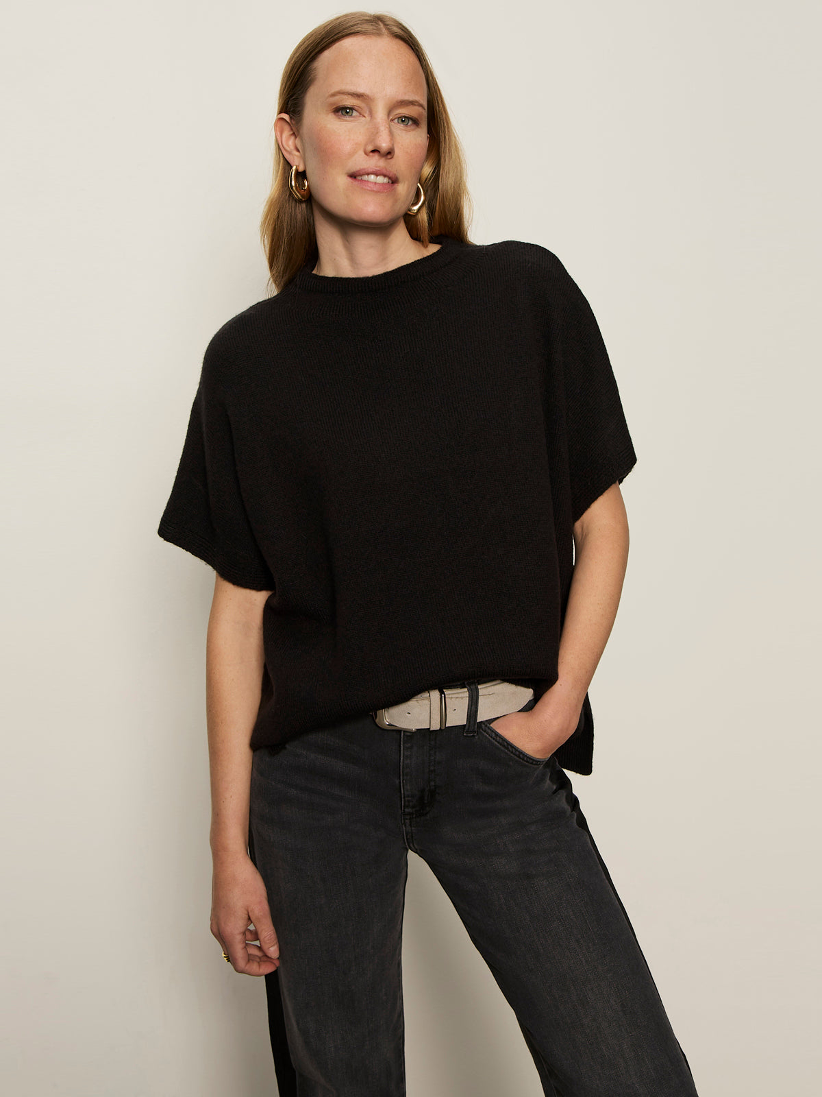 A woman with light skin and straight, light brown hair models the Sanctuary Clothing easy layering shell in black, paired with dark jeans and a light gray belt, standing against a plain background with a relaxed expression.