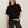 A woman with light skin and straight, light brown hair models the Sanctuary Clothing easy layering shell in black, paired with dark jeans and a light gray belt, standing against a plain background with a relaxed expression.