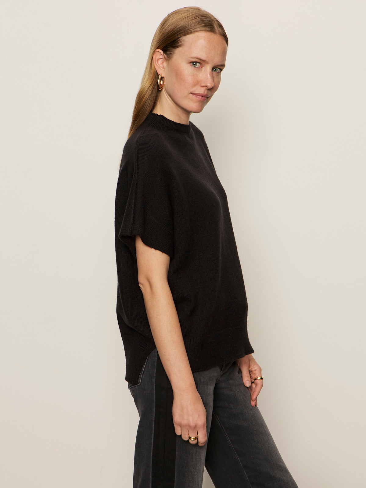 A woman with light brown hair wears the Sanctuary Clothing easy layering shell in black and dark jeans, standing against a plain light wall and looking at the camera with a slight smile.