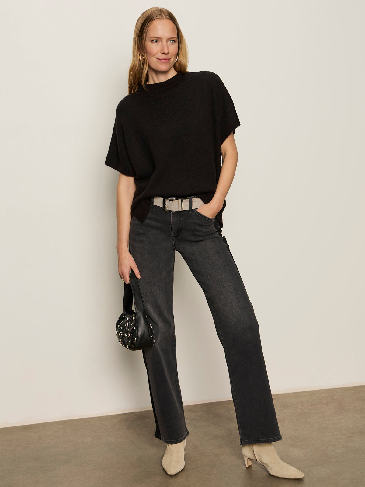 A woman with straight, shoulder-length blonde hair smiles slightly as she stands against a plain background, wearing the Sanctuary Clothing easy layering shell in black. She pairs it with dark gray jeans, beige boots, and a small black handbag.
