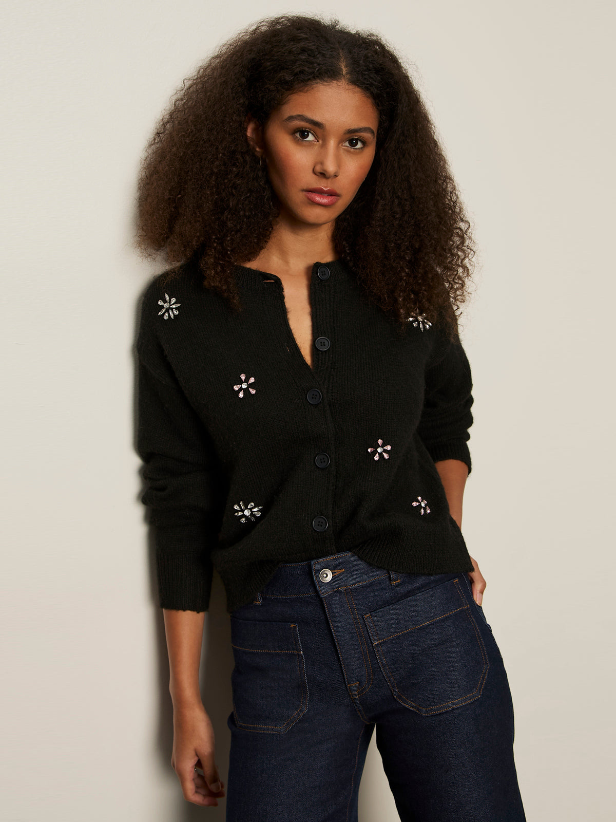 A woman with curly hair models the Sanctuary Clothing must have gem cardi black, a button-up sweater with small floral accents, paired with high-waisted dark blue jeans against a light-colored wall.