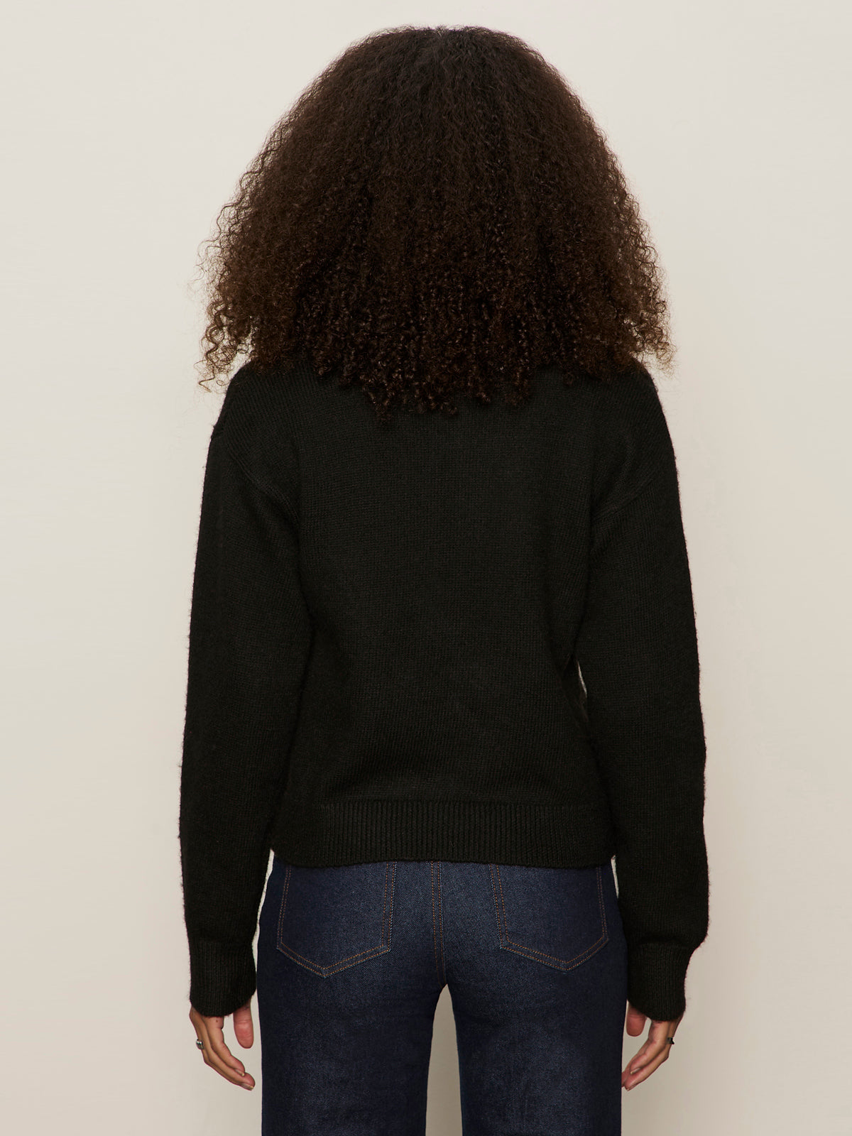 A person with curly brown hair, seen from the back against a plain light background, is wearing Sanctuary Clothing's must have gem cardi in black and dark blue jeans.