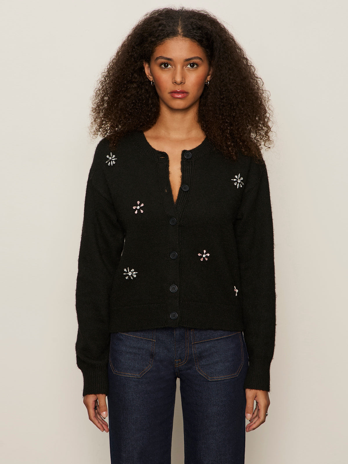 A woman with curly hair wears the Sanctuary Clothing must have gem cardi in black with embroidered florals and dark blue jeans, standing against a plain light background.