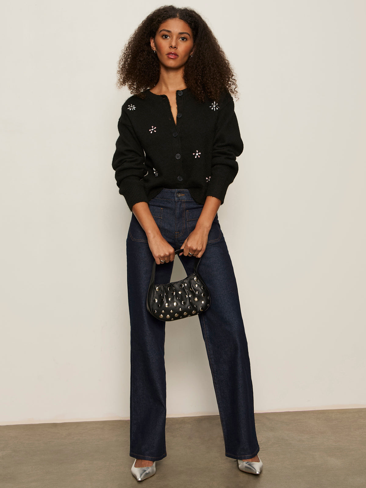 A woman with curly hair wears the Sanctuary Clothing must have gem cardi black, dark blue wide-leg jeans, silver pointed shoes, and holds a black studded handbag against a plain background.