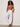 A woman wears Sanctuary Clothing's in the mix sweater tank in ancient clay, paired with white jogger pants and black sandals, standing against an off-white wall with her hands in her pockets.