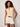 A woman with curly hair wears the Sanctuary Clothing manhattan rib button cardi in banana, paired with white shorts and a brown belt, standing against a light wall and looking at the camera with a relaxed expression.