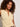 A woman with curly hair stands against a plain light background, wearing the Sanctuary Clothing manhattan rib button cardi banana and looking at the camera with a neutral expression.