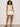 A woman poses against a light background wearing Sanctuary Clothing’s manhattan rib button cardi banana, styled with cream belted shorts, black sandals, and bracelets; she has one hand in her pocket and a relaxed expression.