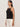 A woman stands in profile against a light background, wearing the Sanctuary Clothing "for keeps pointelle tank" in black with thin straps and off-white trousers. Her straight dark hair is worn down, and she has a neutral expression.