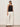 A woman stands against a light wall wearing the Sanctuary Clothing for keeps pointelle tank in black, black sunglasses, wide-leg white pants, and black sandals. Her hands are in her pockets as she looks to the side.