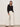 A woman with straight dark hair stands against a plain wall wearing the Sanctuary Clothing for keeps pointelle tank in black, wide-leg white pants, and black sandals. She poses relaxed with one hand in her pocket and a neutral expression.