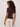 A woman with curly hair and sunglasses wears the Sanctuary Clothing "perfect knitwear tee rich soil," a short-sleeved brown ribbed top, paired with light pants, as she stands looking over her shoulder against a plain background.