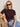 A woman with blonde hair and sunglasses sits confidently on a woven chair, wearing the Sanctuary Clothing perfect knitwear tee in rich soil and blue jeans, accessorized with large bangles, against a neutral background.