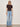 A woman in Sanctuary Clothing's perfect knitwear tee in rich soil, paired with light blue high-waisted jeans, sandals, a brown belt, sunglasses, and chunky bracelets stands against a plain light-colored wall.