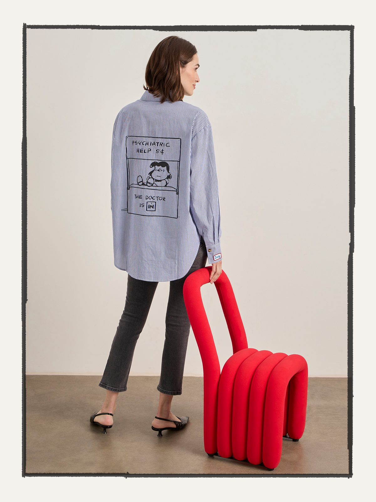 A person wearing the Sanctuary Clothing "the doctor is in shirt blue and white stripe" holds a bold red wavy chair indoors, styled with black jeans and heels.