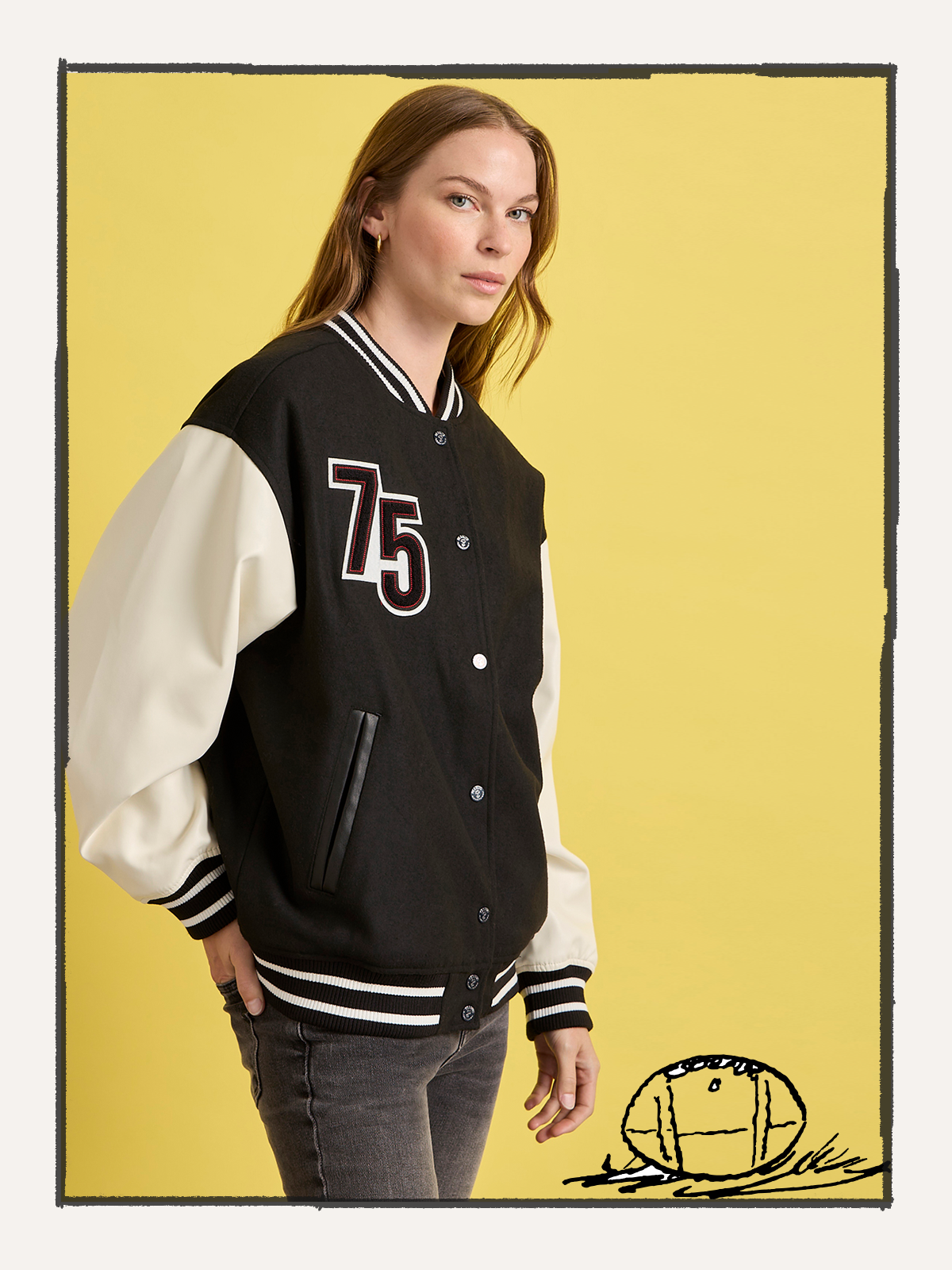 A woman with long hair models the Sanctuary Clothing 75 varsity jacket in black and white, standing before a yellow background with a small football helmet graphic in the bottom right corner.