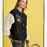 A woman with long hair models the Sanctuary Clothing 75 varsity jacket in black and white, standing before a yellow background with a small football helmet graphic in the bottom right corner.