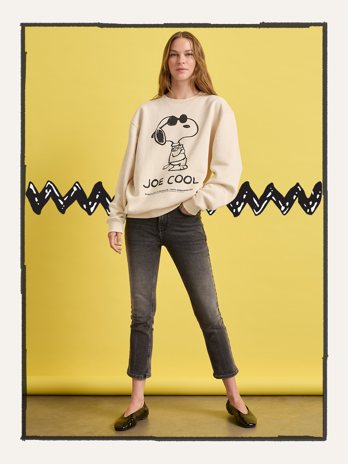 Against a yellow backdrop with a black zigzag pattern, a person wears Sanctuary Clothing's "Joe Cool Sweatshirt Collagen," paired with gray jeans and black shoes.
