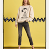 Against a yellow backdrop with a black zigzag pattern, a person wears Sanctuary Clothing's "Joe Cool Sweatshirt Collagen," paired with gray jeans and black shoes.