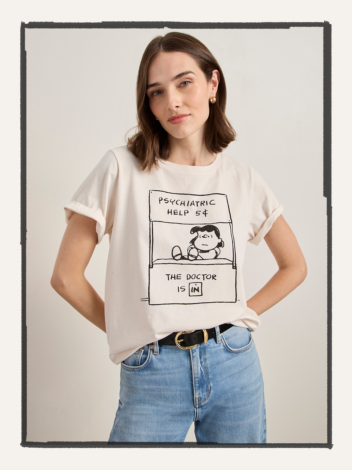 A woman stands with her hands on her hips, wearing light blue jeans and the Sanctuary Clothing "the doctor is in tee collagen," a white T-shirt with a Peanuts cartoon of Lucy at her "Psychiatric Help 5¢. The Doctor is IN" booth.