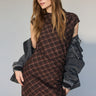 A woman with long brown hair wears the Sanctuary Clothing autumns best sweater dress in cocoa plaid, featuring a thigh-high slit, styled with a black denim jacket over her shoulders, posing against a neutral background.