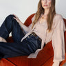 A woman with long brown hair and bangs sits on an orange velvet chair, wearing the Sanctuary Clothing "Fade Into You" ruffle blouse in fawn and high-waisted dark blue jeans, looking at the camera with a relaxed expression.