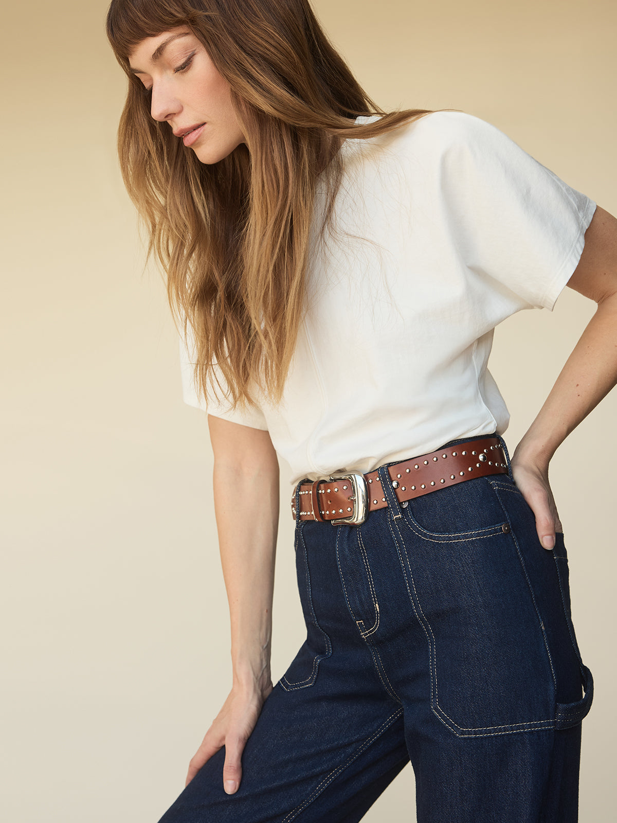 A woman with long brown hair wears the Sanctuary Clothing shirred waist dolman tee in white, tucked into high-waisted dark blue jeans with visible stitching and a brown studded belt, standing against a beige background.