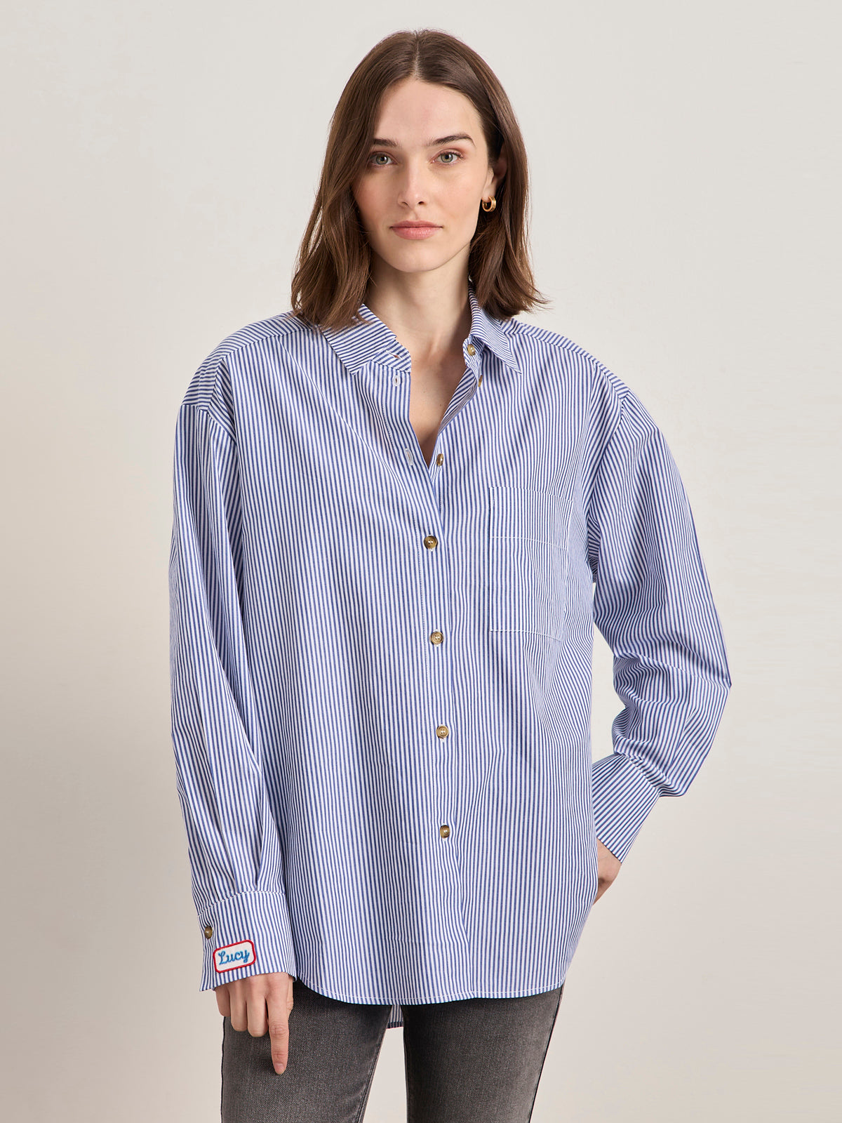 A woman with shoulder-length brown hair wears the Sanctuary Clothing x PEANUTS 75th Anniversary "the doctor is in" blue and white stripe shirt, featuring gold buttons, styled with gray jeans, against a plain light background.