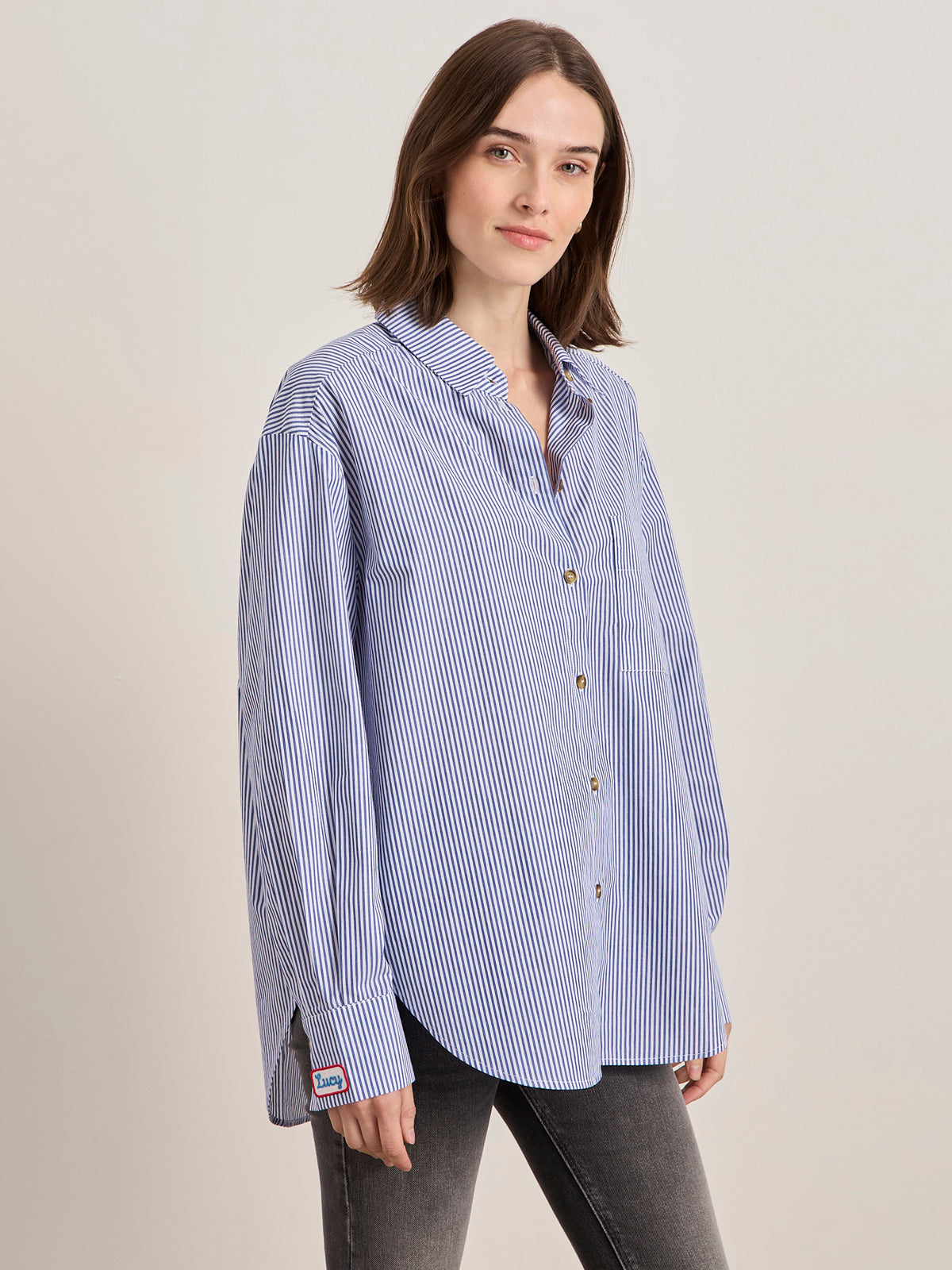 A woman with shoulder-length brown hair models the Sanctuary Clothing x PEANUTS 75th Anniversary "the doctor is in" blue and white stripe shirt against a plain light background.