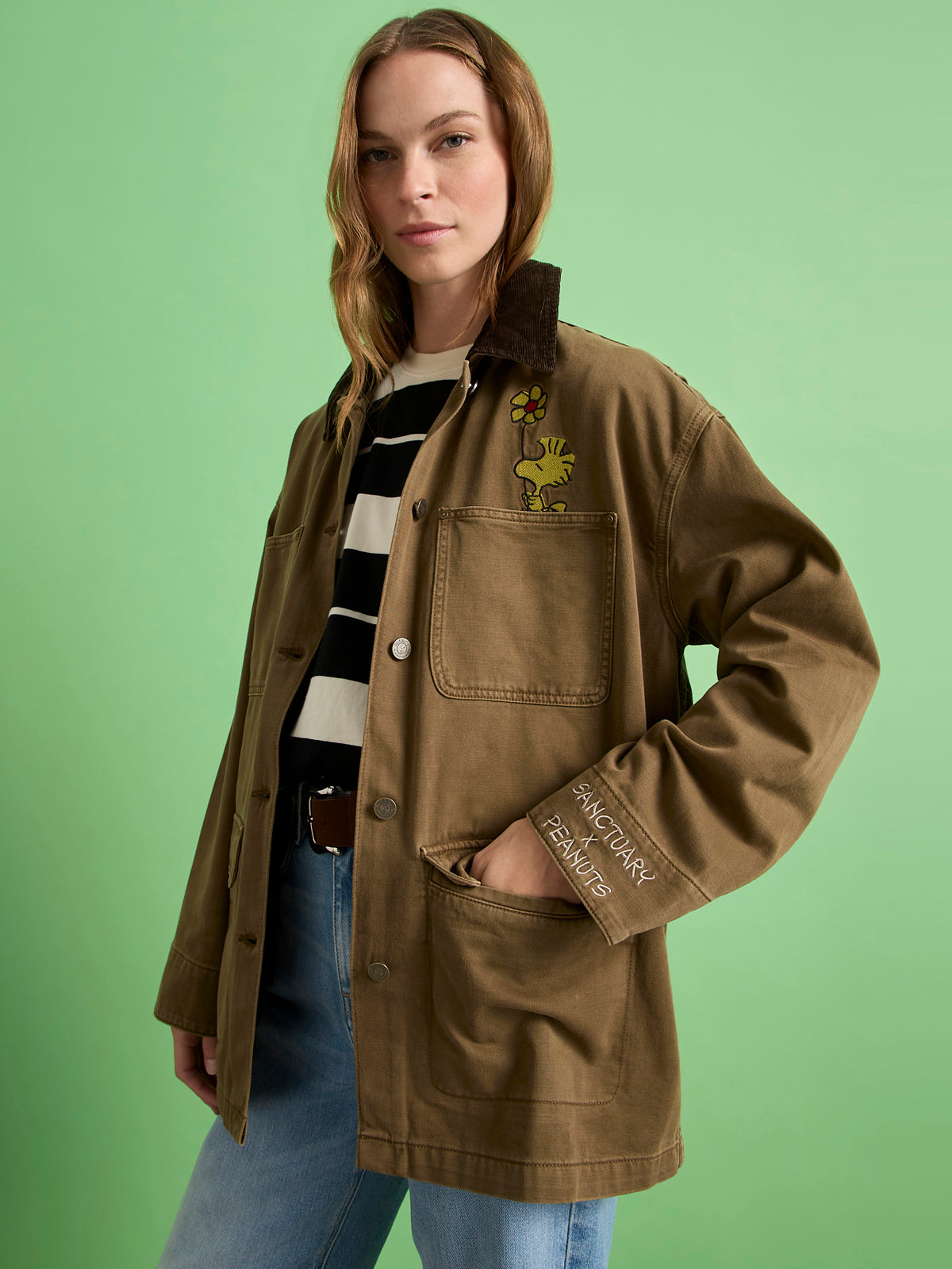 A woman stands confidently against a green background wearing the Sanctuary Clothing x PEANUTS 75th Anniversary save our planet jacket timber, paired with a black-and-white striped shirt and blue jeans.