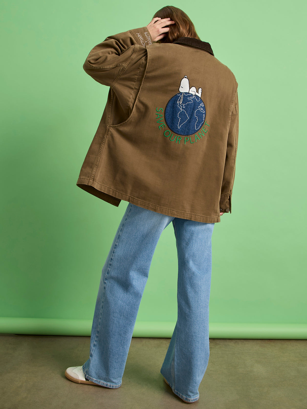 A person stands with their back to the camera, wearing the save our planet jacket timber by Sanctuary Clothing x PEANUTS 75th Anniversary, featuring Snoopy and Woodstock on a globe, paired with light blue jeans and cream shoes.