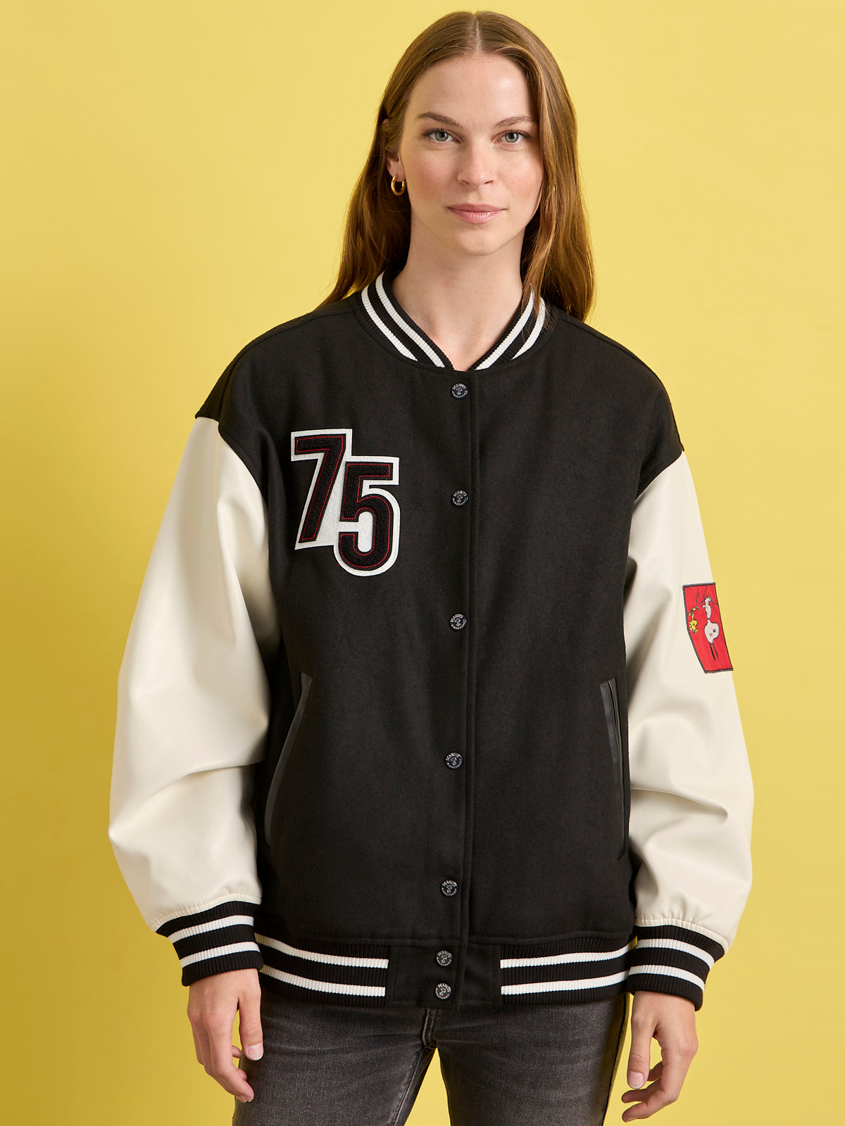 A woman models the Sanctuary Clothing x PEANUTS 75th Anniversary "75 varsity jacket black white" with a red sleeve patch, standing against a yellow background and smiling slightly with her hands in her pockets.