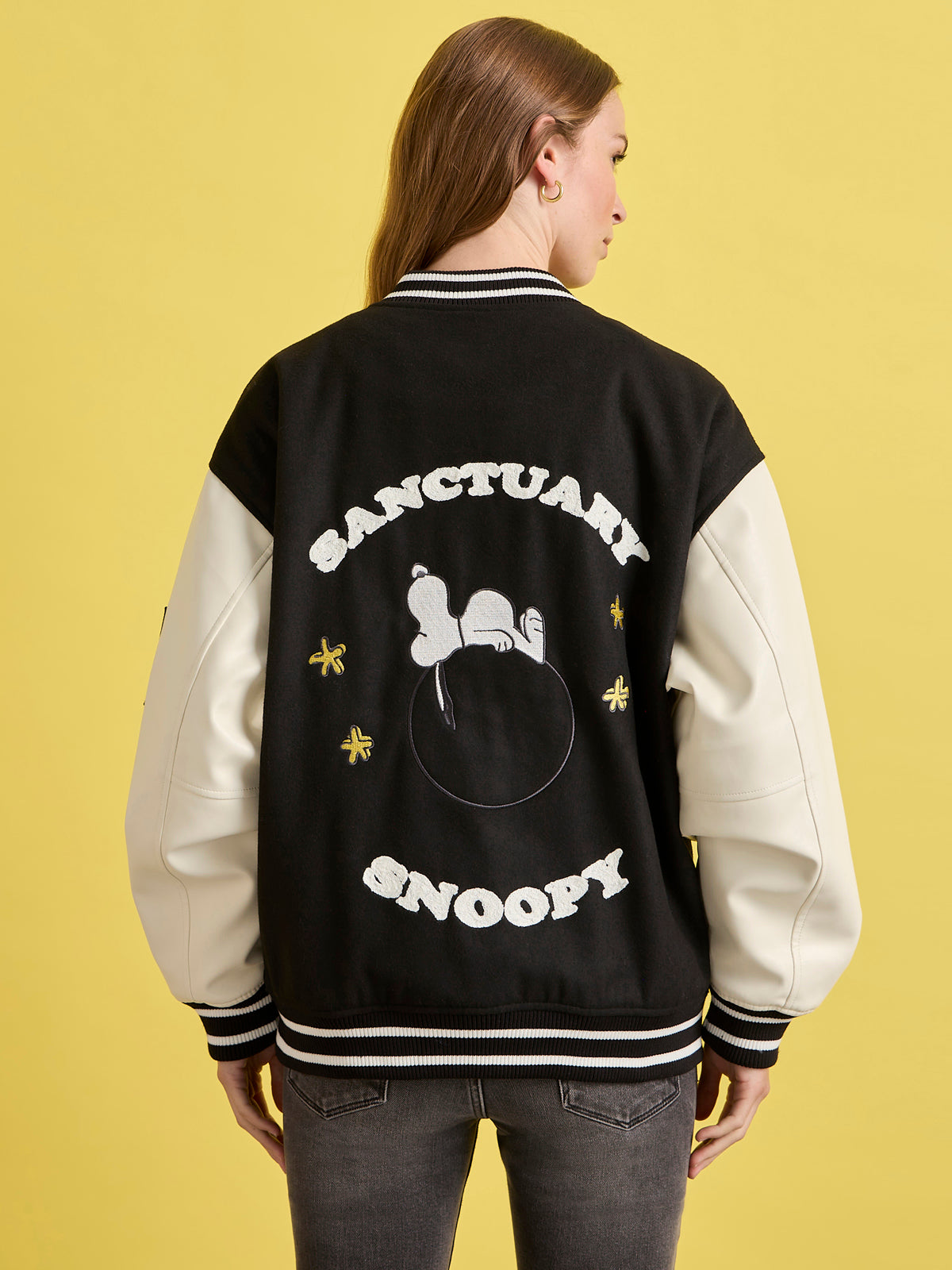 A person with long hair wears the Sanctuary Clothing x PEANUTS 75th Anniversary 75 varsity jacket in black and white, featuring Snoopy on the back with yellow stars and "SANCTUARY SNOOPY" written above.