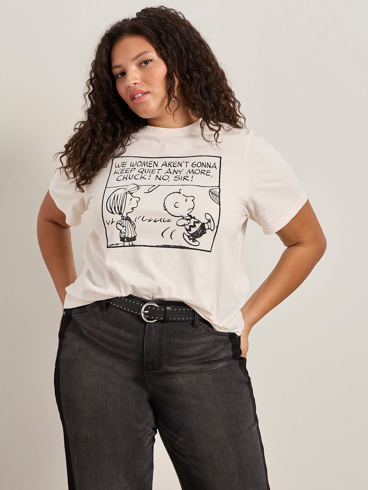 A woman with curly hair wears the Sanctuary Clothing x PEANUTS 75th Anniversary "arent gonna keep quiet tee pink milk" and black jeans, standing with one hand in her pocket against a plain light background.
