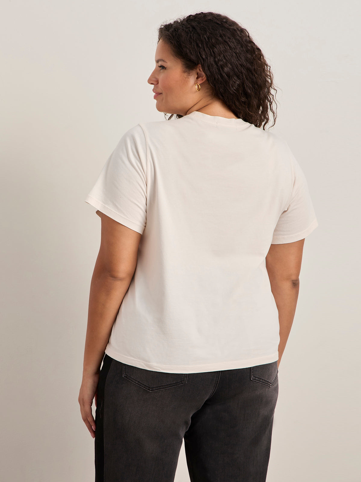 A woman with curly hair faces away, wearing the "arent gonna keep quiet tee pink milk" by Sanctuary Clothing x PEANUTS 75th Anniversary and dark jeans, against a light background.