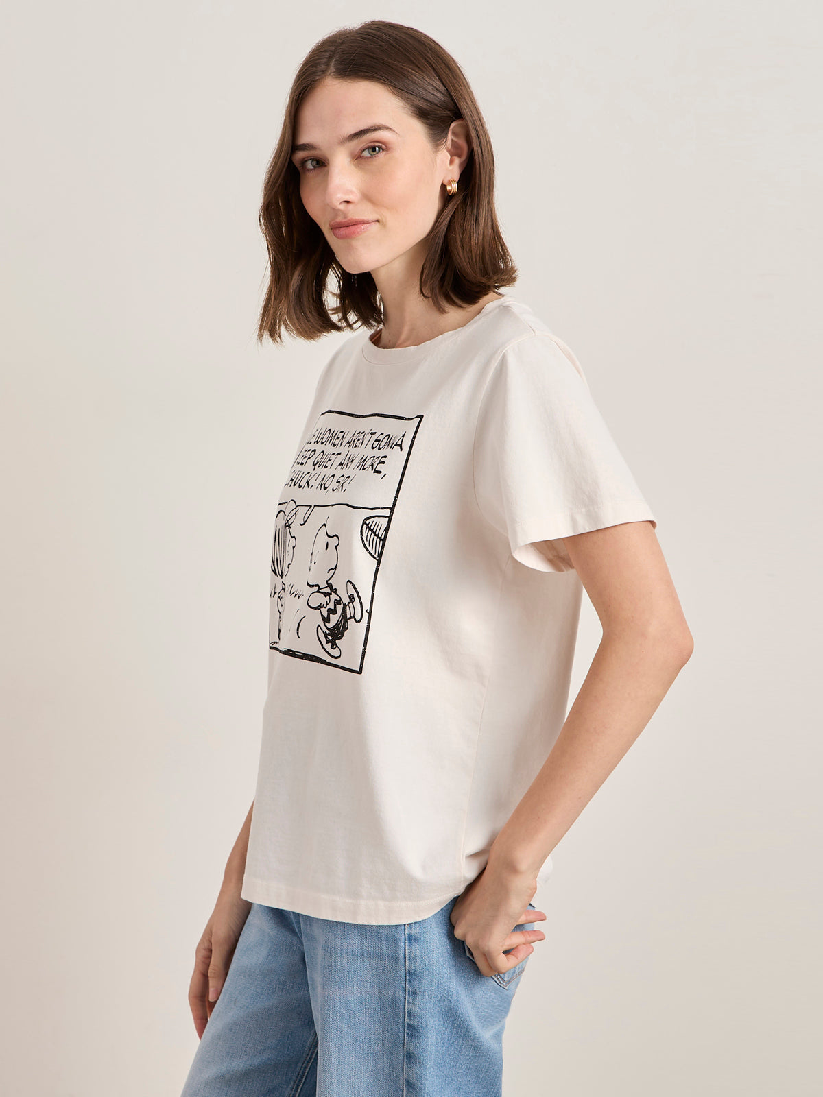 A woman with shoulder-length brown hair wears the Sanctuary Clothing x PEANUTS 75th Anniversary "arent gonna keep quiet tee pink milk" and light blue jeans, standing against a plain light background.