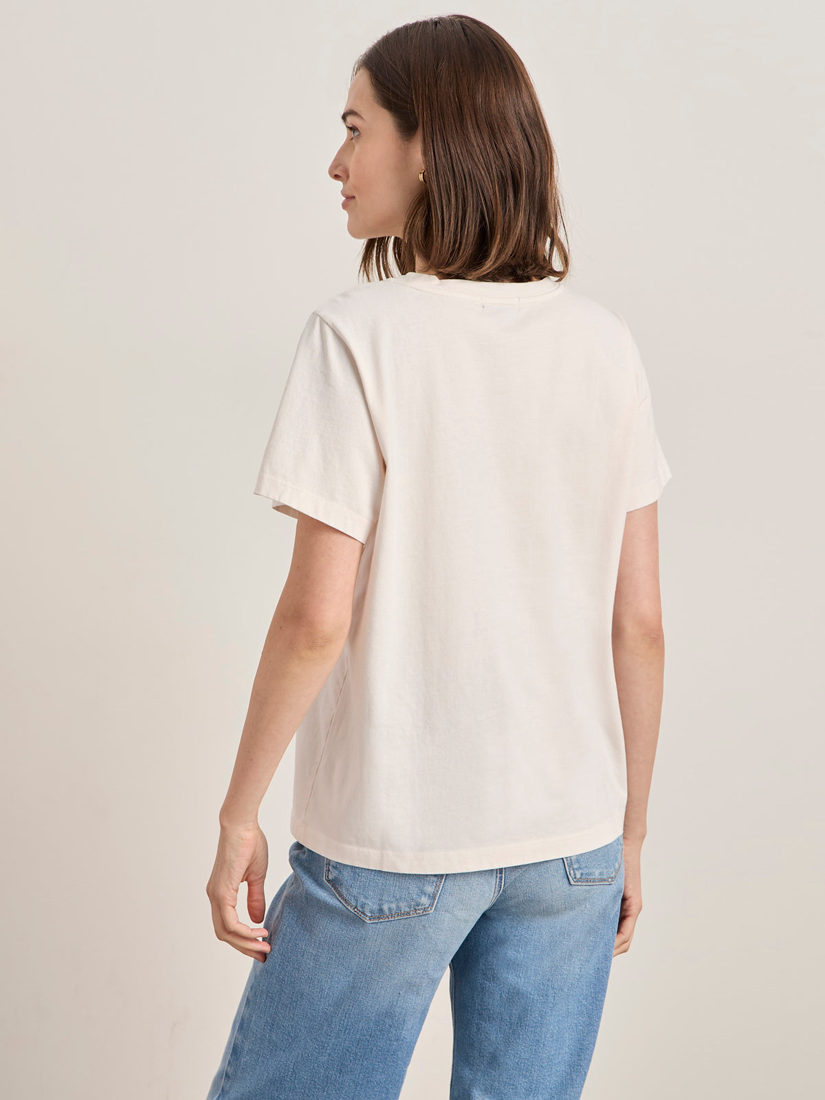 A woman with straight brown hair wears the arent gonna keep quiet tee in pink milk by Sanctuary Clothing x PEANUTS 75th Anniversary, paired with blue jeans, standing facing away from the camera against a neutral background.